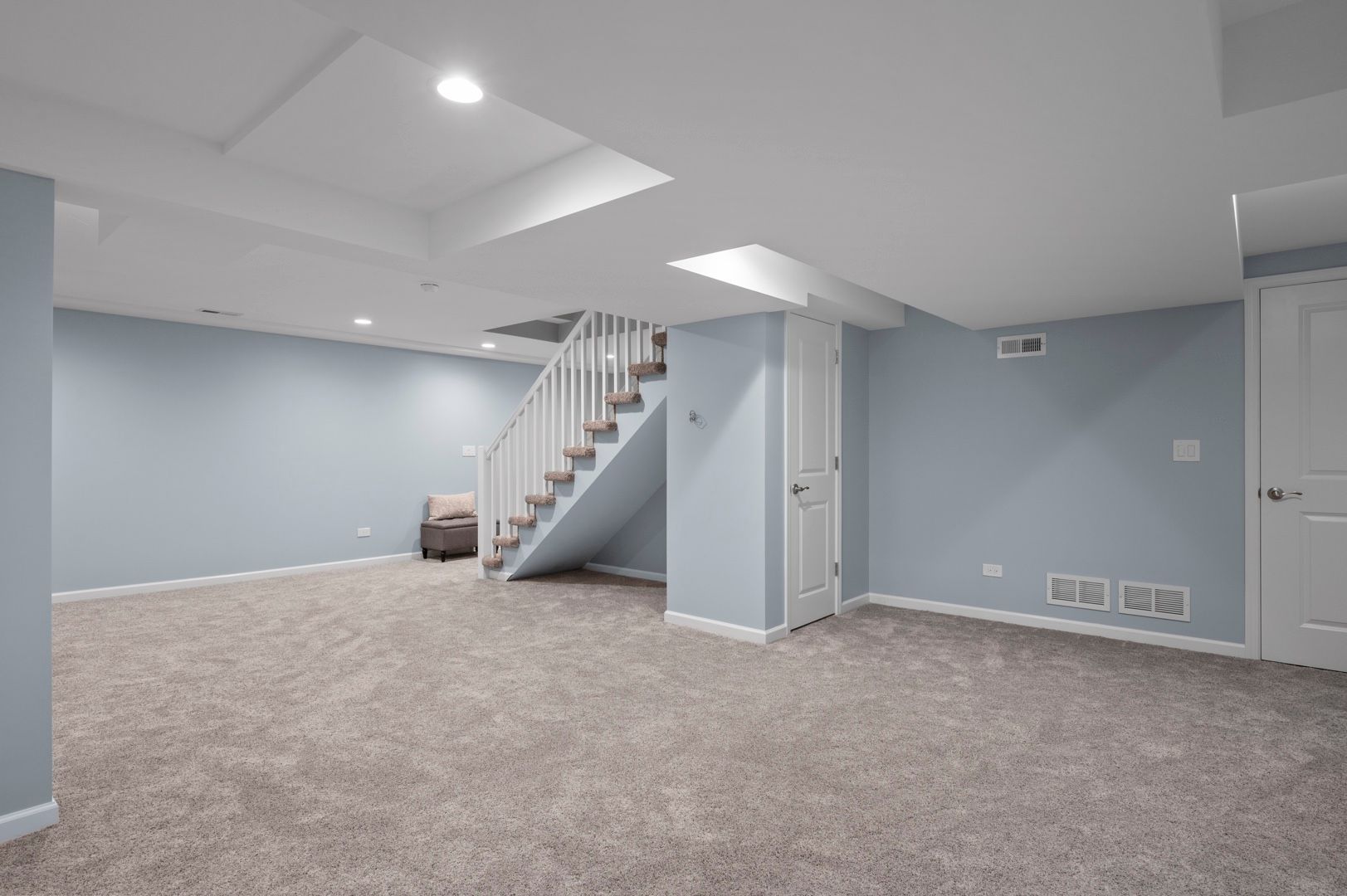 Spacious, light blue basement room with stairs, white door and trim, and gray carpet.