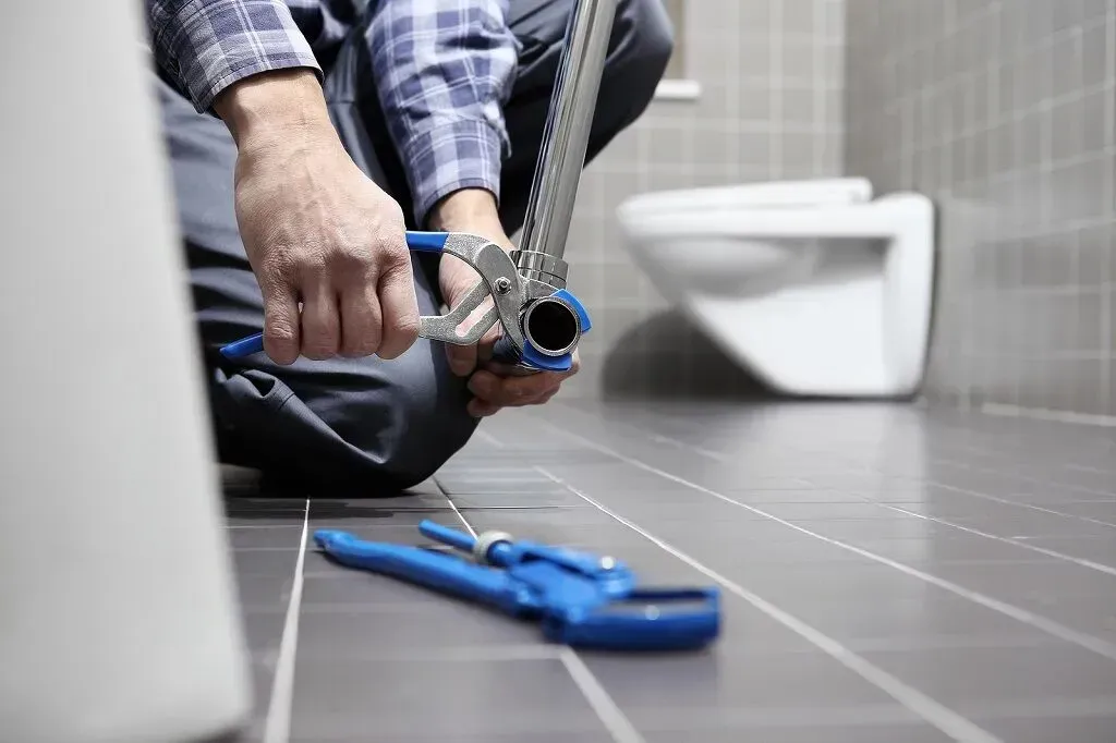 Plumber using a wrench to work on a pipe in a bathroom with a white toilet.