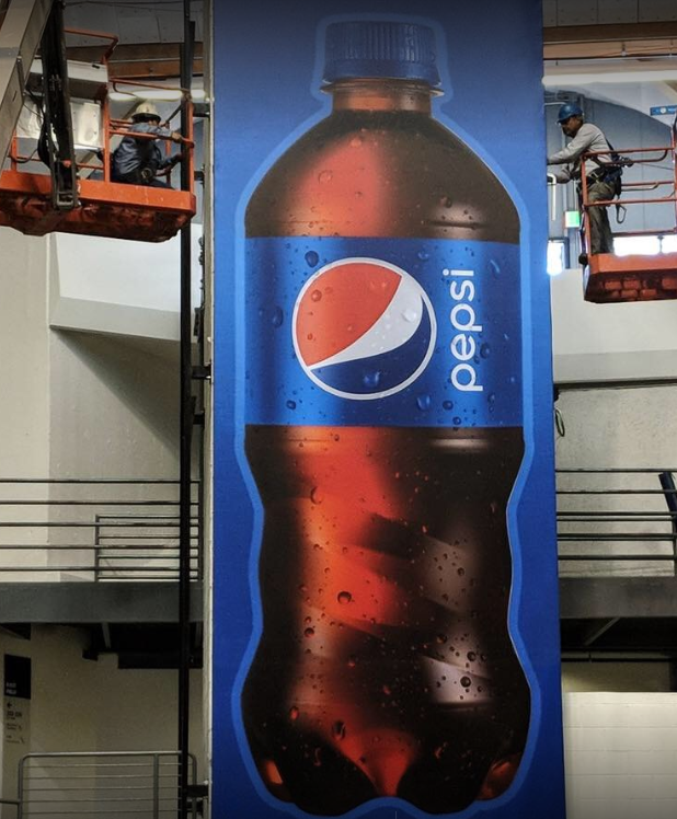 Workers install a large Pepsi bottle advertisement indoors.