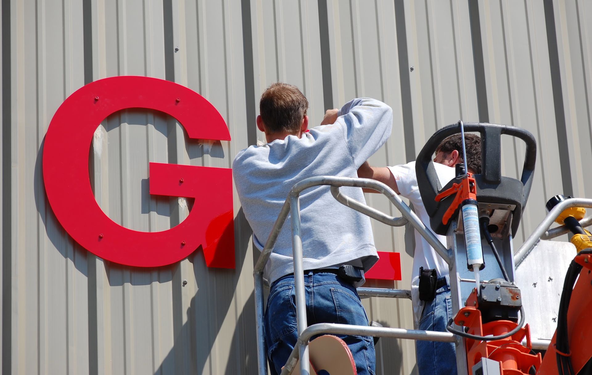 Workers installing large red channel-letter signage on a metal building wall.