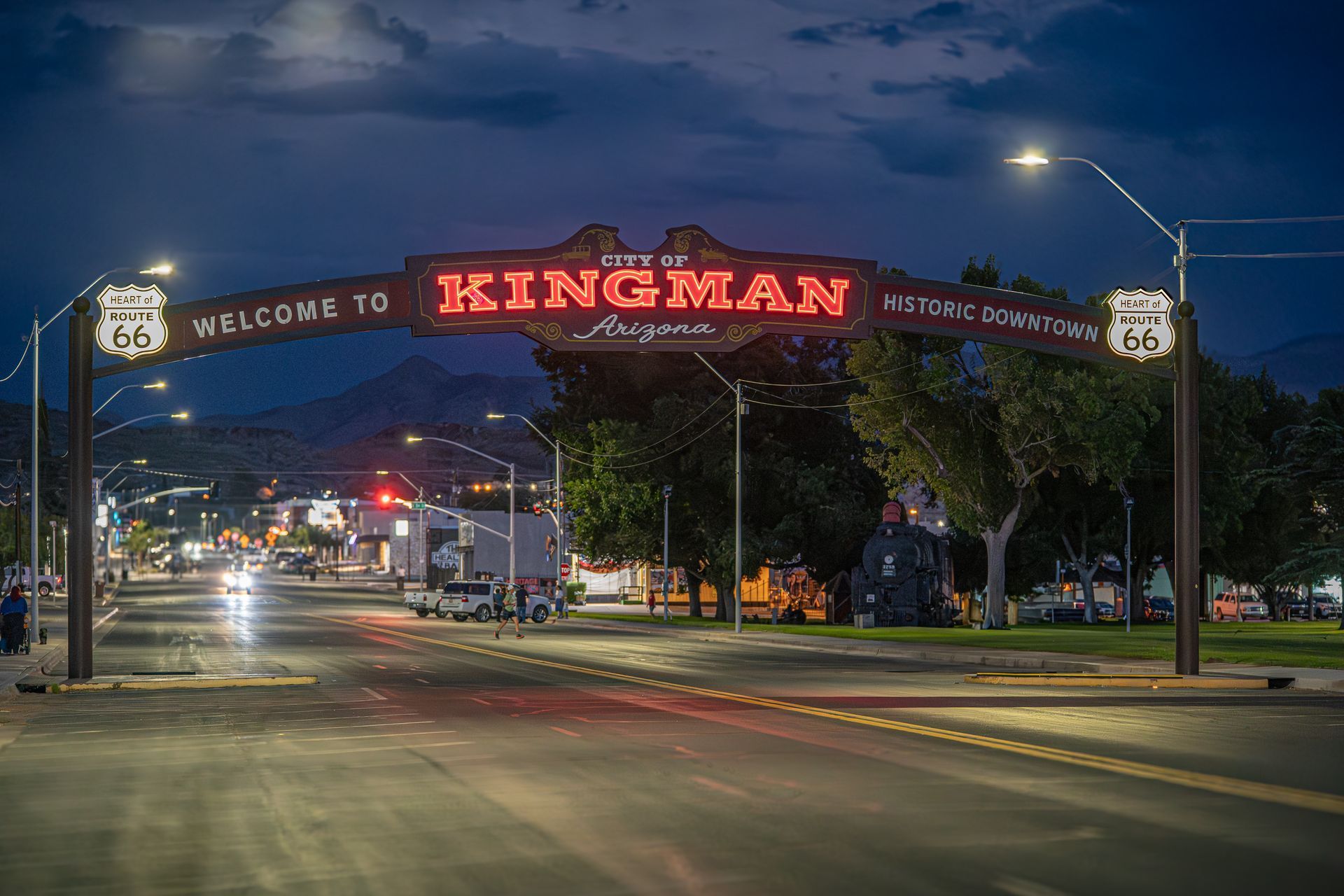 Welcome arch to Kingman, Arizona, lit at night. Route 66 signage. Cars on street, mountains in background.