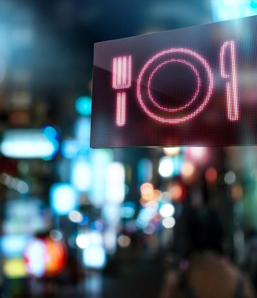 Neon restaurant sign with fork, plate, and knife against blurred city lights.