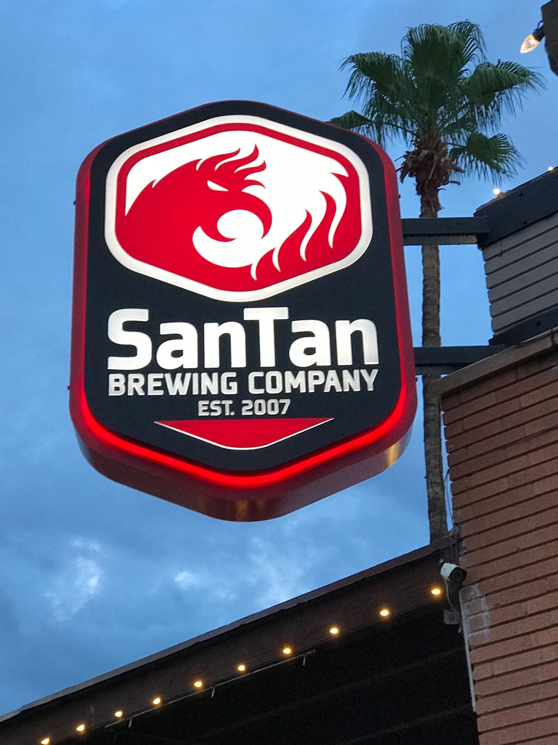 Sign for SanTan Brewing Company, red and white logo on black background. Palm tree and twilight sky in background.