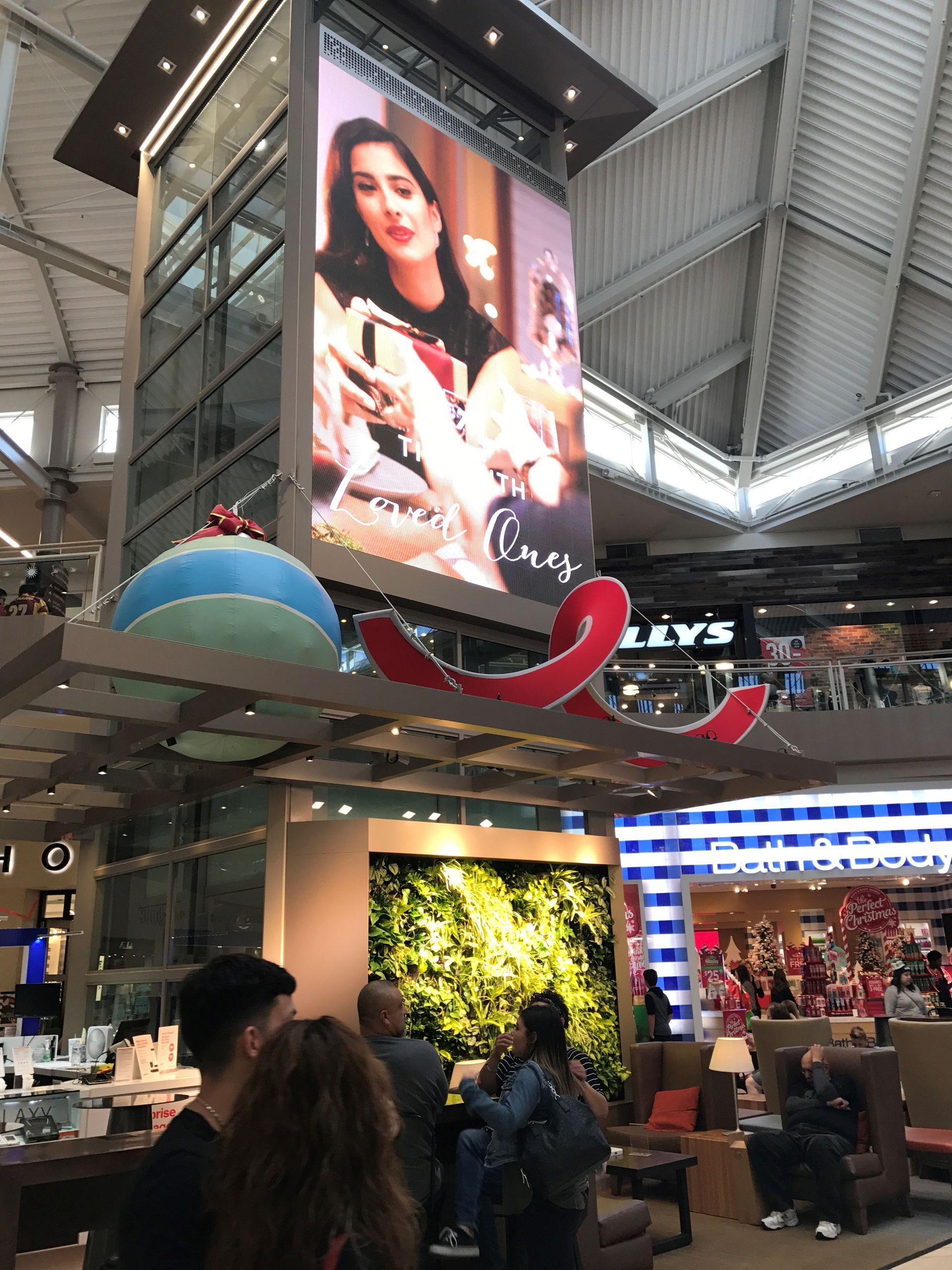Large digital display in a mall shows a woman with text, over store with a green wall and a seating area.