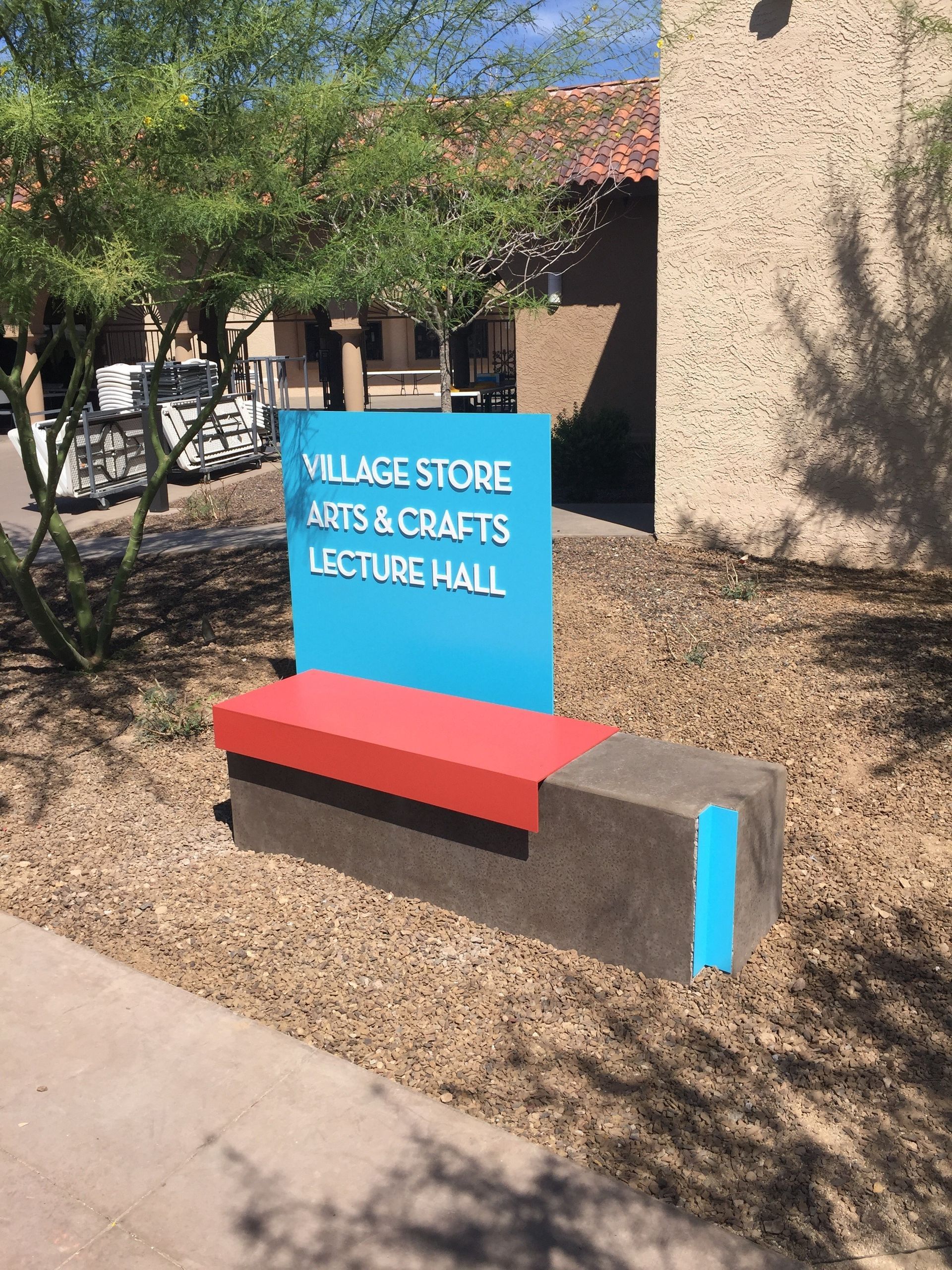 Sign for Village Store Arts & Crafts Lecture Hall with a red bench and turquoise accents.