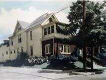 A historic light-colored house with a dark brown lower level, angled rooflines, and a large tree in the front yard.