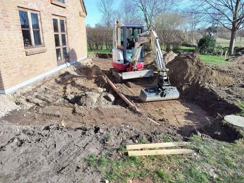 En lille gravemaskine graver et hul i snavset foran et murstenshus.