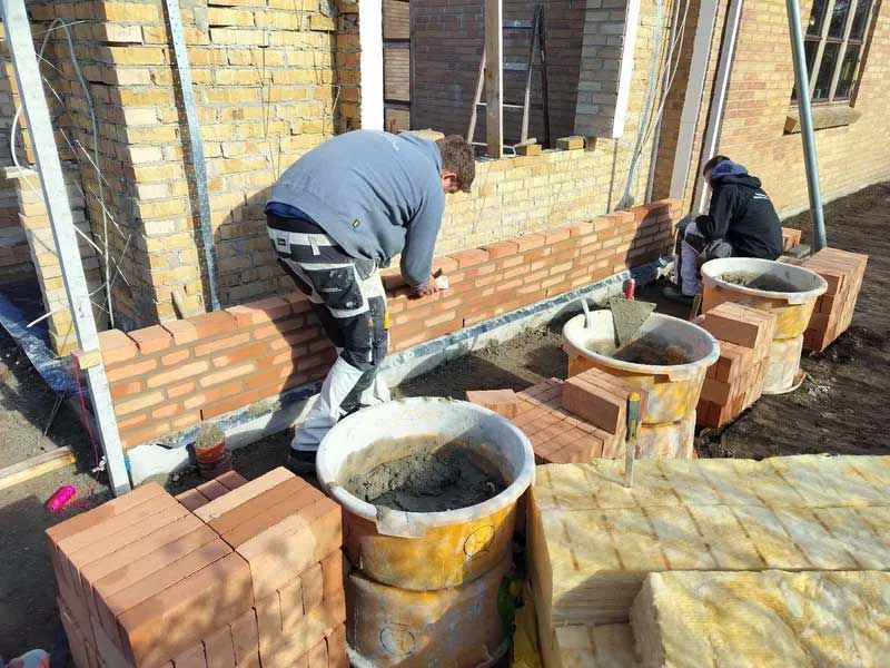 Murere bygger en murstensvæg udendørs. En lægger mursten, en anden blander mørtel. Sollys.