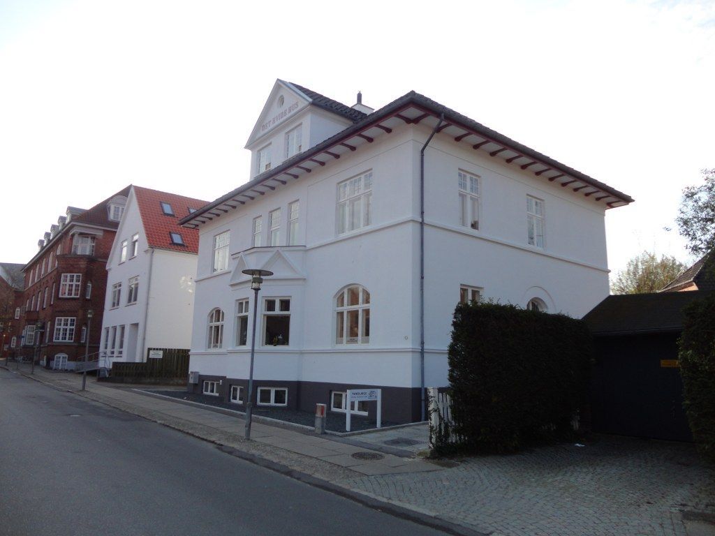 En stor hvid bygning med rødt tag ligger på hjørnet af en gade.