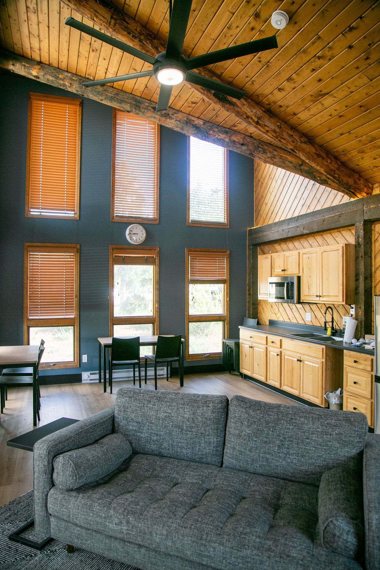 Cozy living room with vaulted wood ceiling, gray sofa, tall windows, and kitchenette area