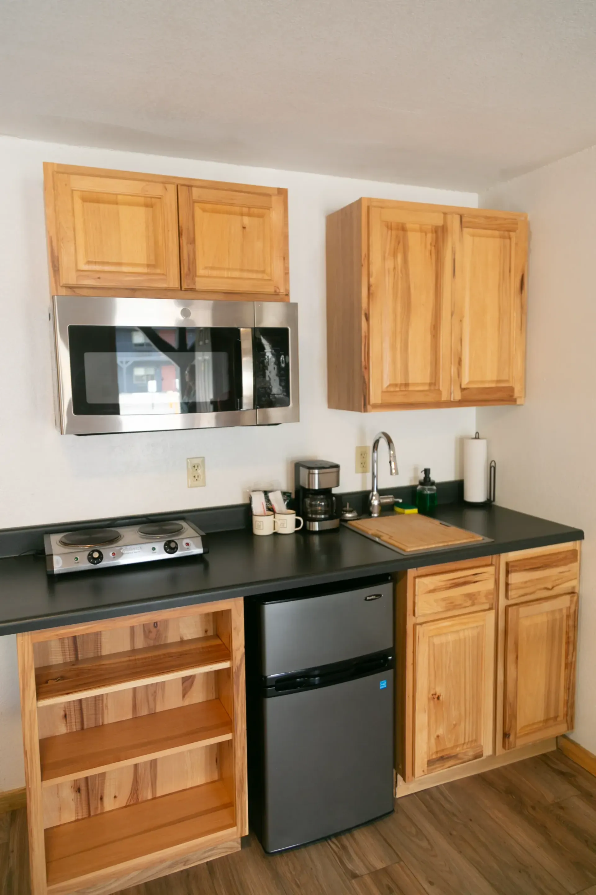 A small kitchenette with light wood cabinets, a stainless steel microwave, a mini-fridge, and a small electric stove.