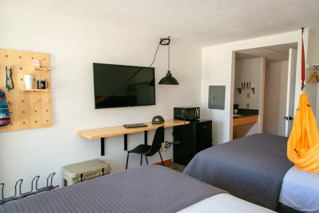 Modern bedroom with two beds, wall-mounted TV, desk, lamp, and orange bag on chair