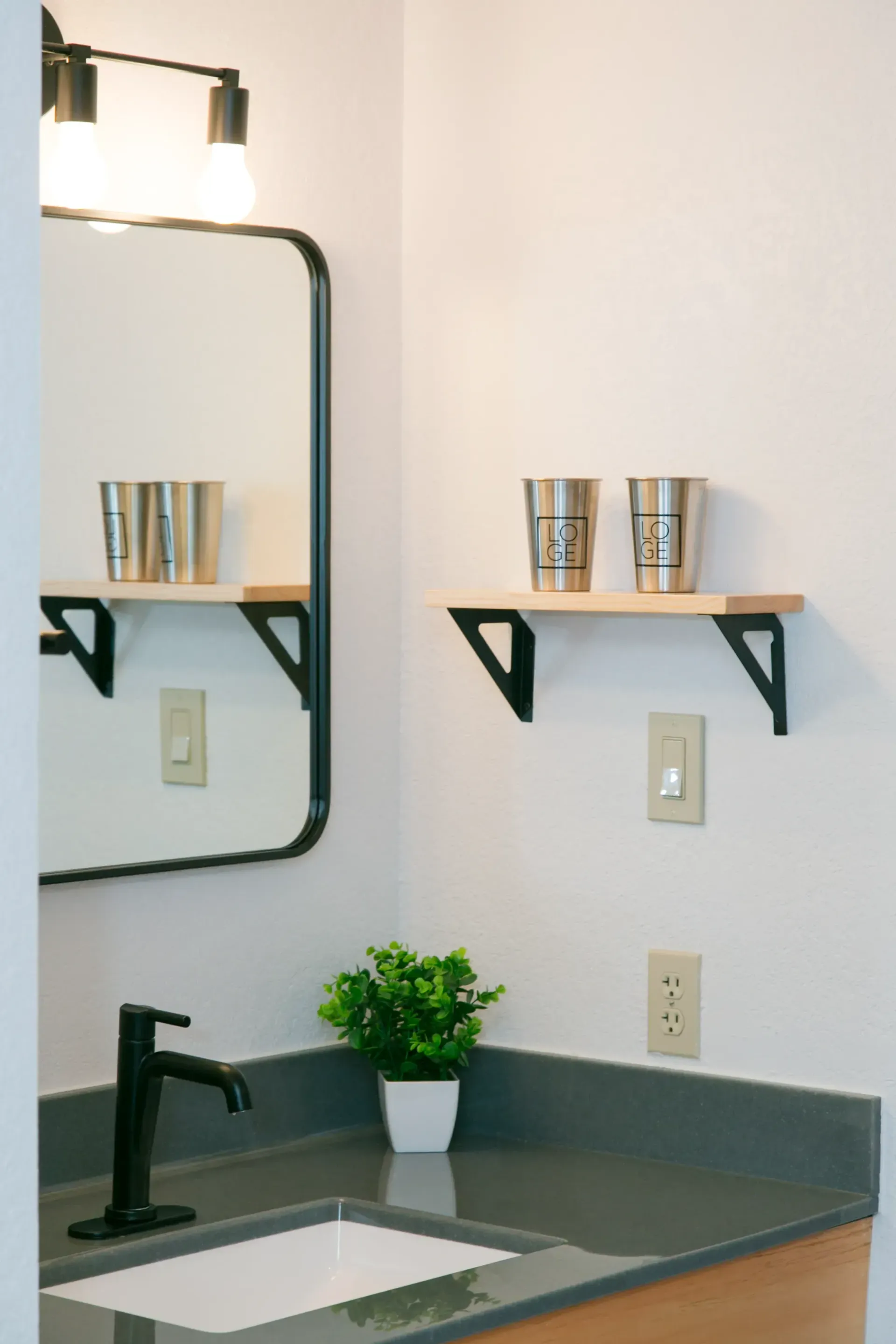 A modern bathroom vanity with a black faucet, rectangular mirror, two floating wood shelves, and a small potted plant.