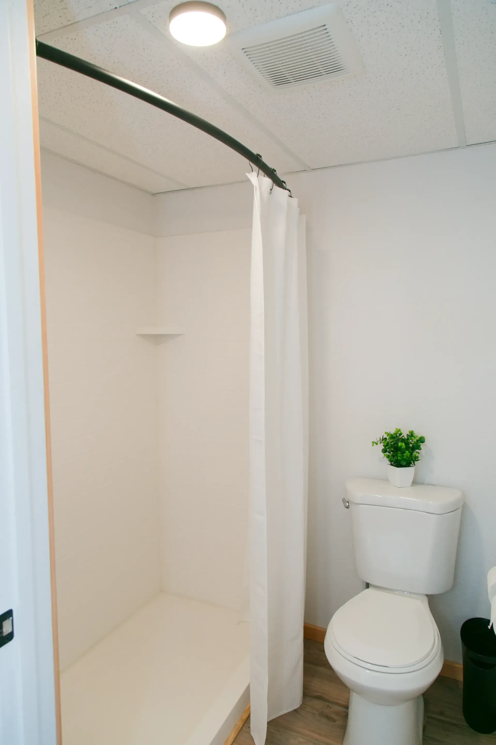 A white bathroom featuring a walk-in shower with a curved rod and white curtain, and a white toilet with a small plant.