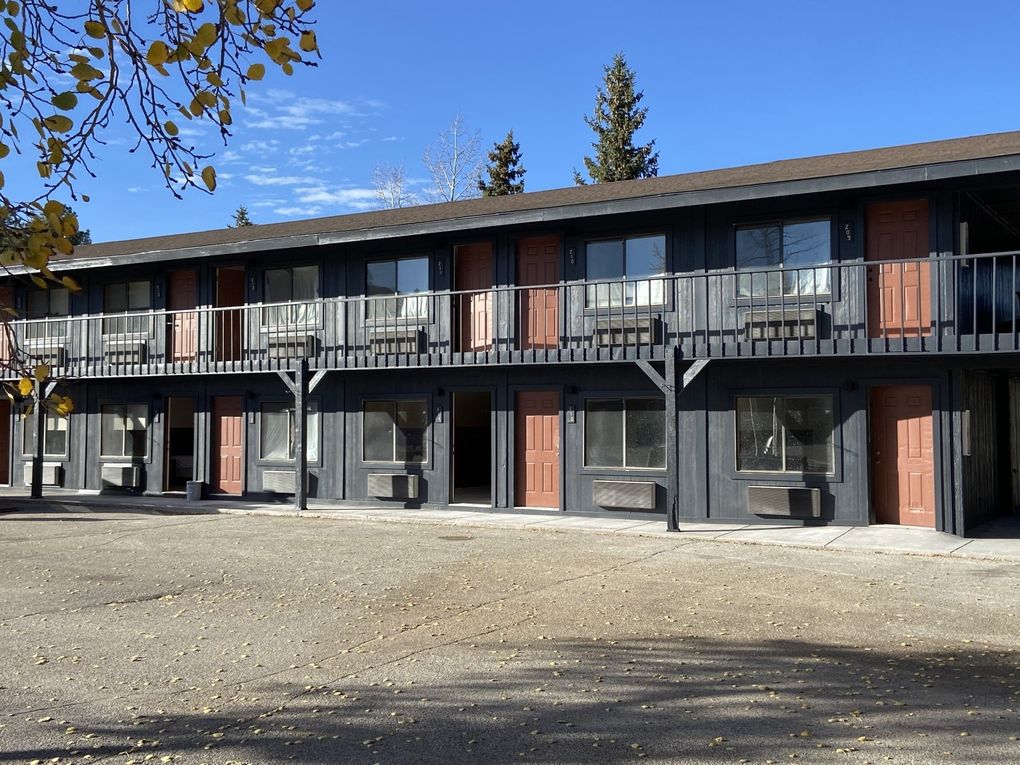 A two-story dark gray motel building with brown doors, a wooden balcony, and a gravel parking lot under a blue sky.