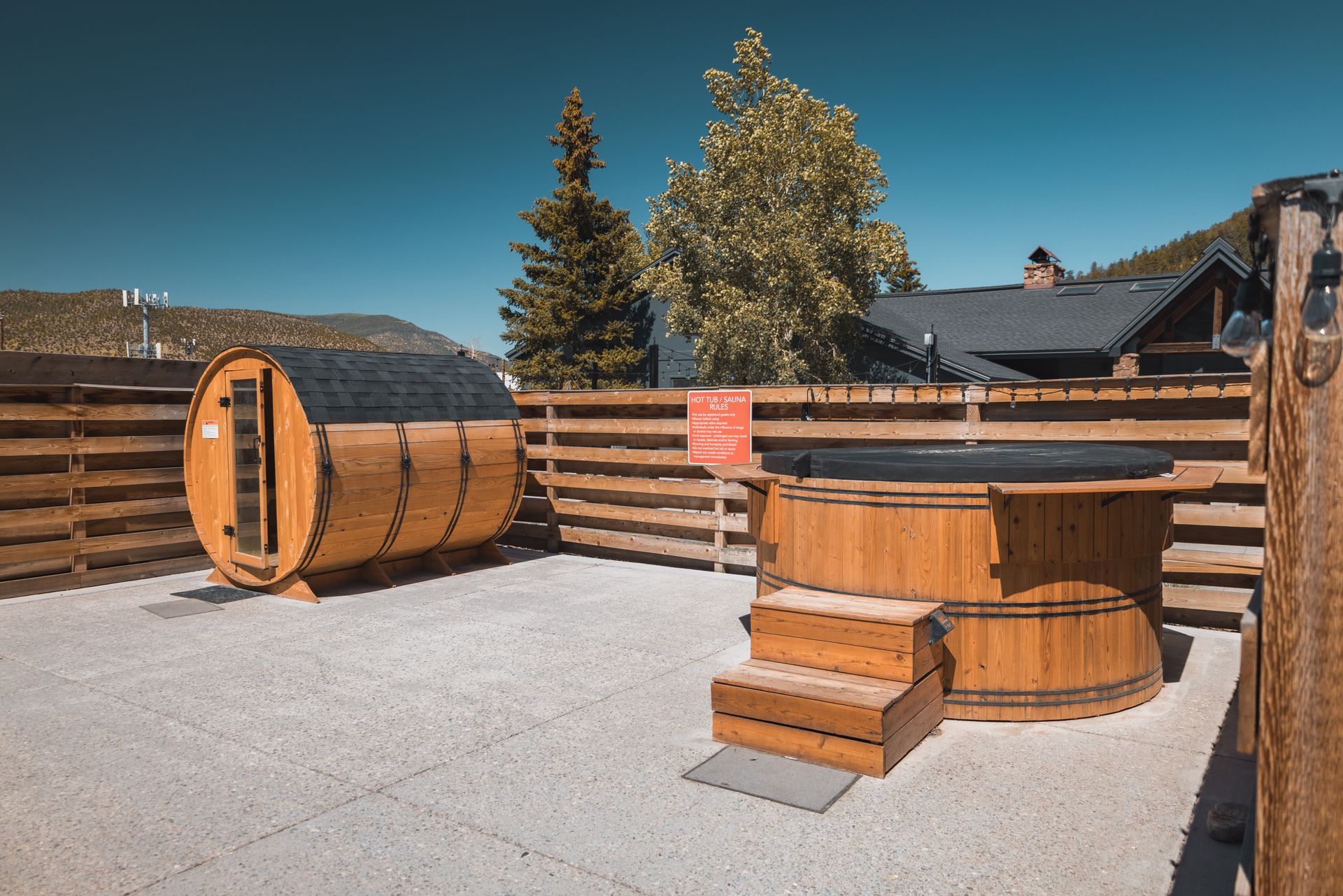 Wooden outdoor sauna barrels and hot tubs on a gravel patio by a fence and trees.