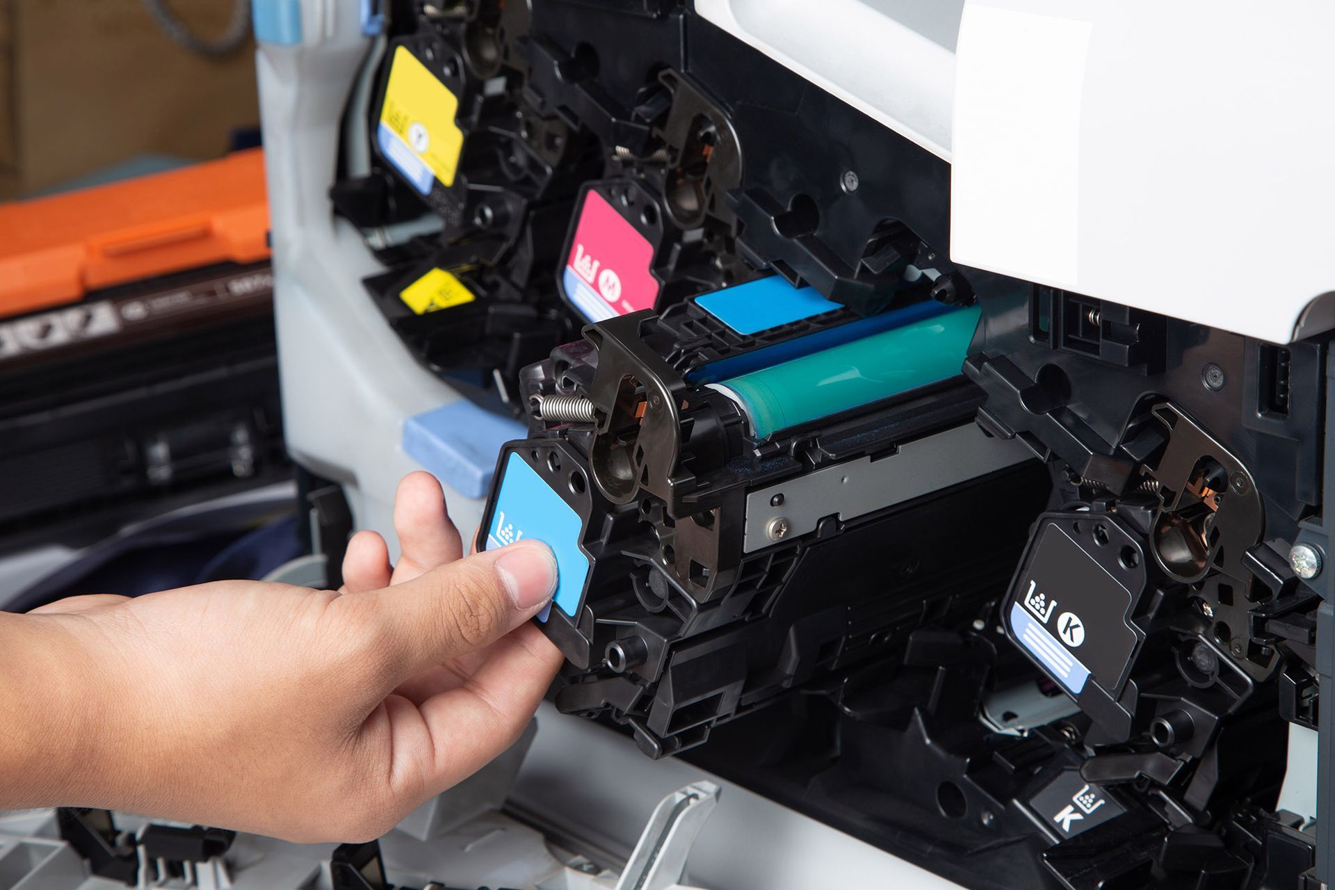 Person replacing a cyan toner cartridge in a color printer; black, cyan, magenta, and yellow cartridges visible. Person replacing a cyan toner cartridge in a color printer; black, cyan, magenta, and yellow cartridges visible.