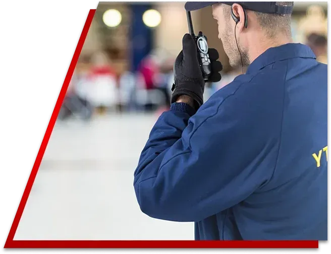 Security guard in blue uniform speaking into a handheld radio, seen from behind against a blurred indoor background.