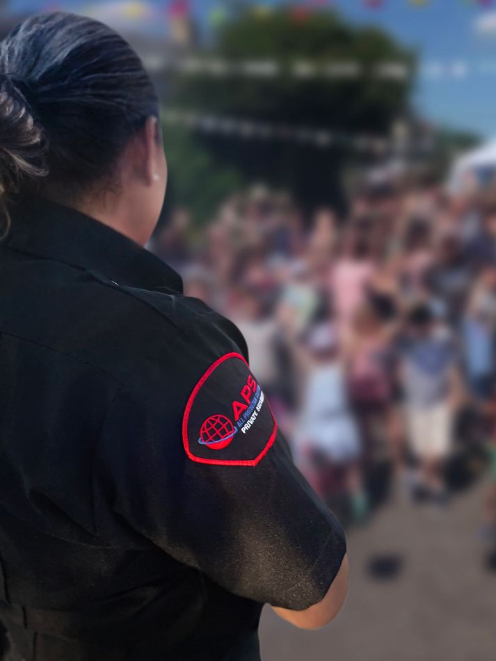 A security guard in a blue uniform speaking into a two-way radio, seen from behind in a blurred, indoor public space.