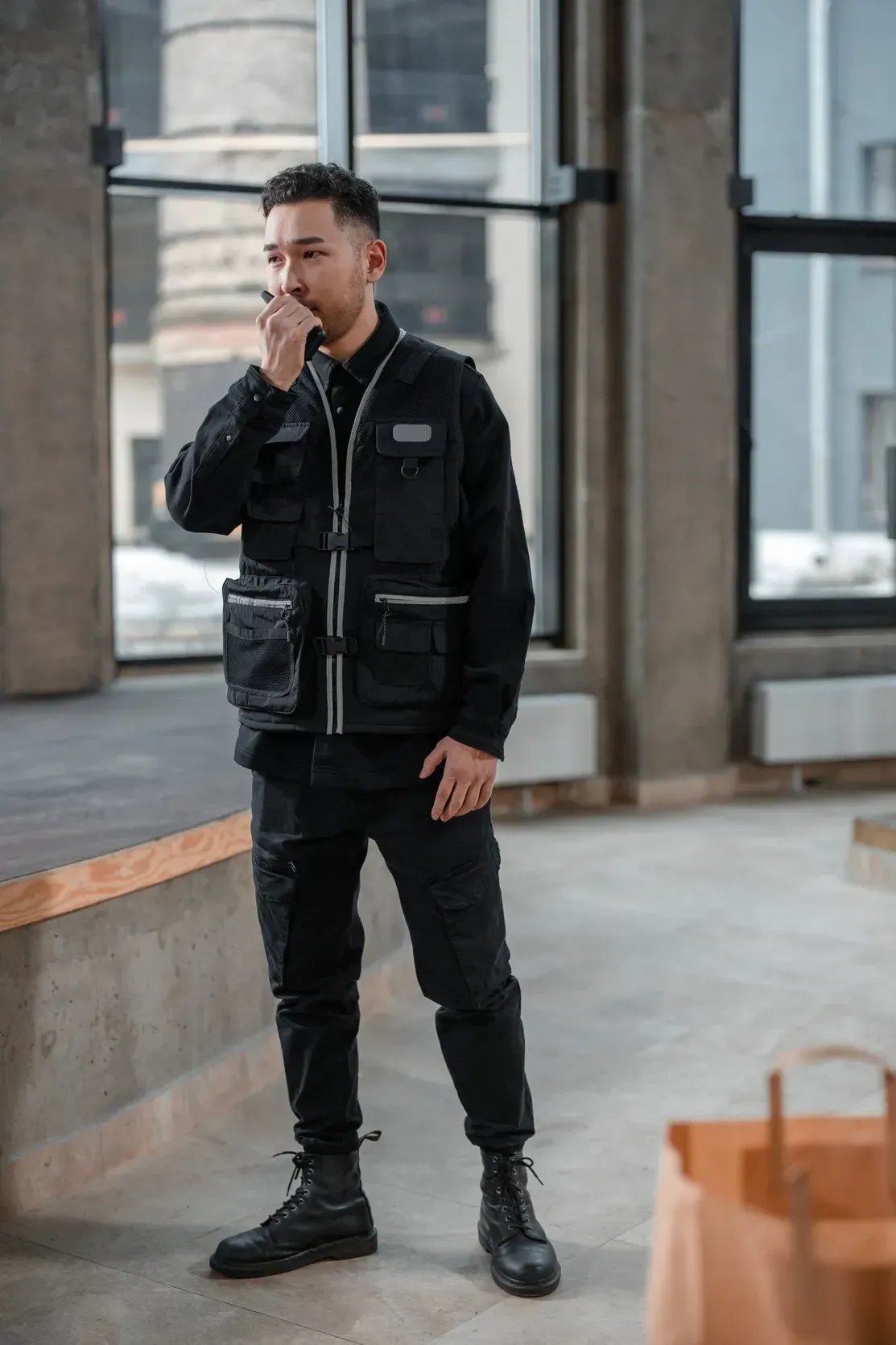 Man in black clothing standing indoors, holding a hand to his face near large windows.