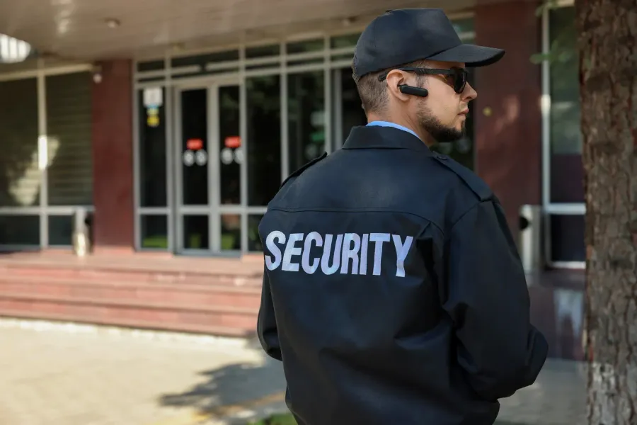 A security guard with arms crossed stands in front of a security patrol vehicle in an apartment complex parking lot.