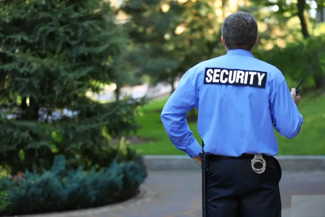 A security guard in a black uniform holding a flashlight, standing on an elevated walkway overlooking a parking area.