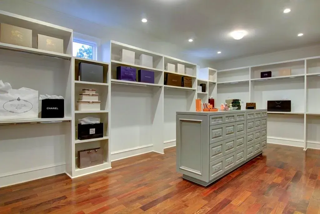 Walk-in closet with white shelves and storage boxes, hardwood floor, grey island, bright lighting.