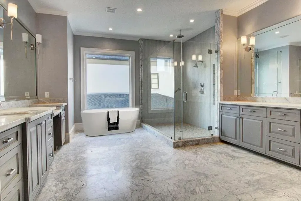 Spacious modern bathroom with freestanding tub, glass shower, dual sinks, gray cabinets, and light gray tile flooring.