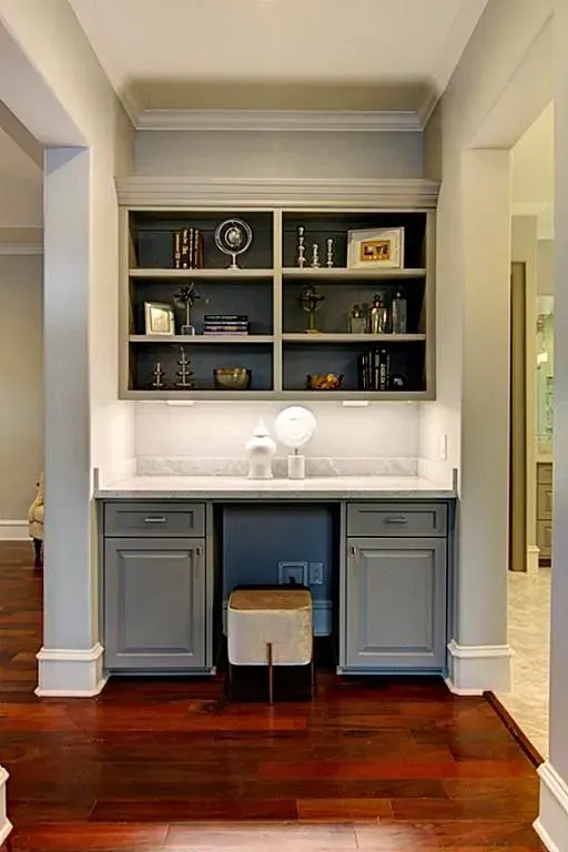 Built-in gray desk with cabinets, marble countertop, and shelves filled with decor. Beige stool sits below.