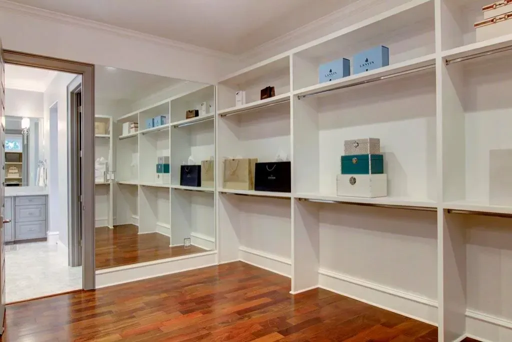 Walk-in closet with white shelves and wooden floor; a mirror reflects the space.