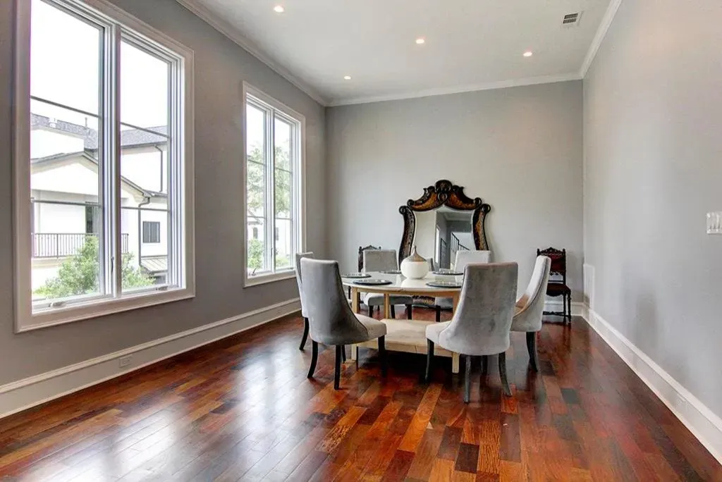 Dining room with round table, six gray chairs, ornate mirror, and large windows. Hardwood floor and gray walls.