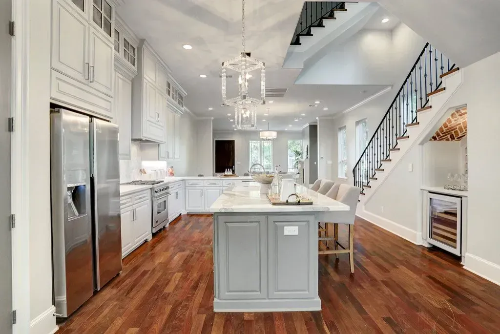Kitchen with white cabinets, stainless steel appliances, a gray island, and a staircase. Hardwood floors.