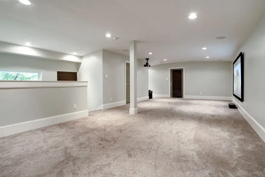 Spacious carpeted room with light gray walls, white trim, recessed lighting, and a dark framed artwork.