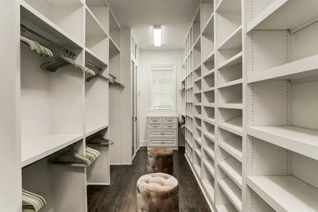 White walk-in closet with shelves and hanging rods. Two upholstered stools sit on a dark wood floor.