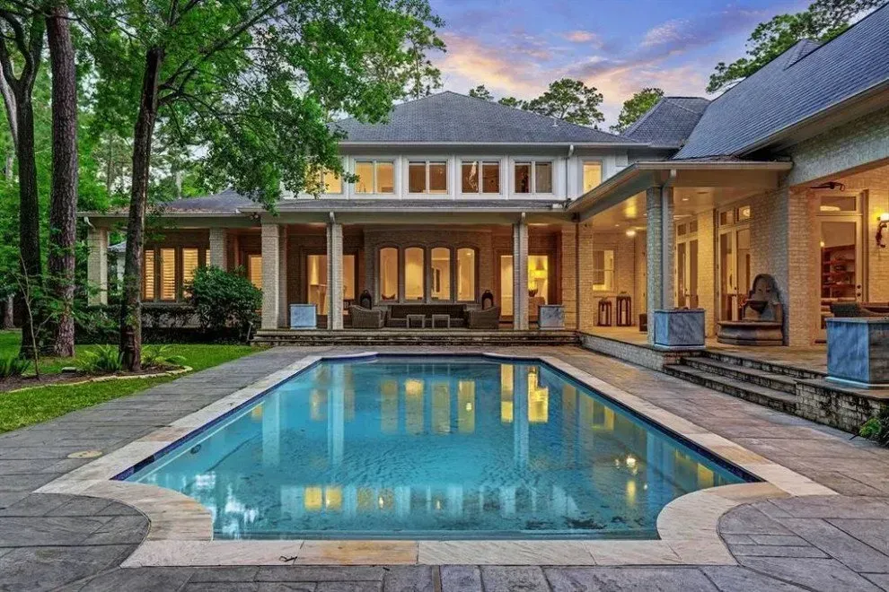 Luxury home exterior with a swimming pool, covered patio, and lush trees.