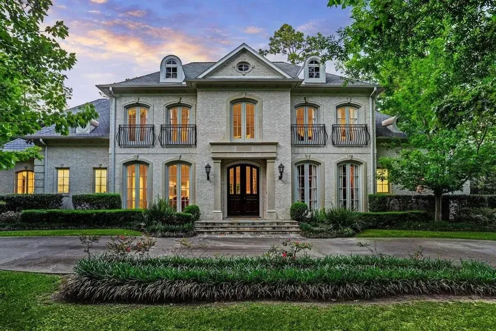 Large, two-story, light-colored brick home with black front door, balconies, and manicured landscaping.