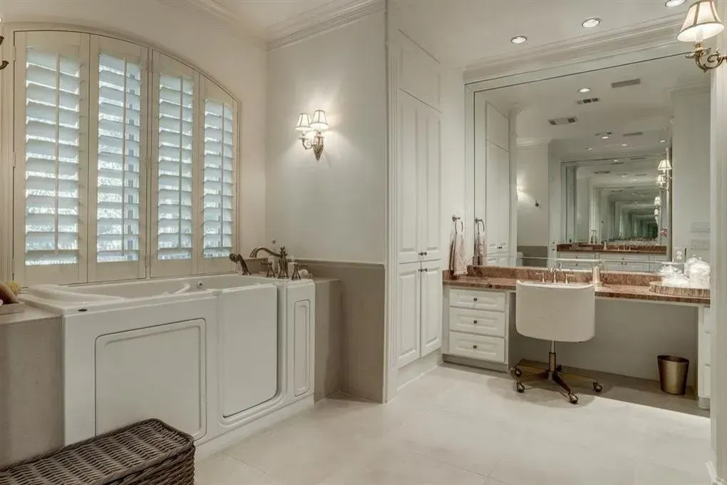 Elegant white bathroom with a walk-in tub, shutters, and a vanity area with a chair.