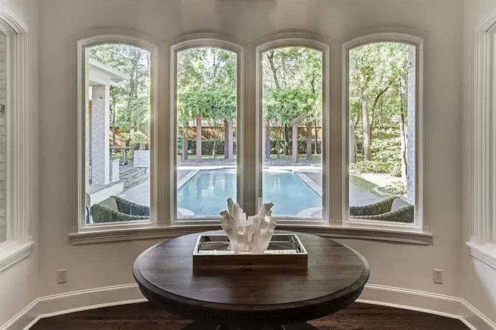 Round wooden table with decor in front of large windows overlooking a pool and trees.