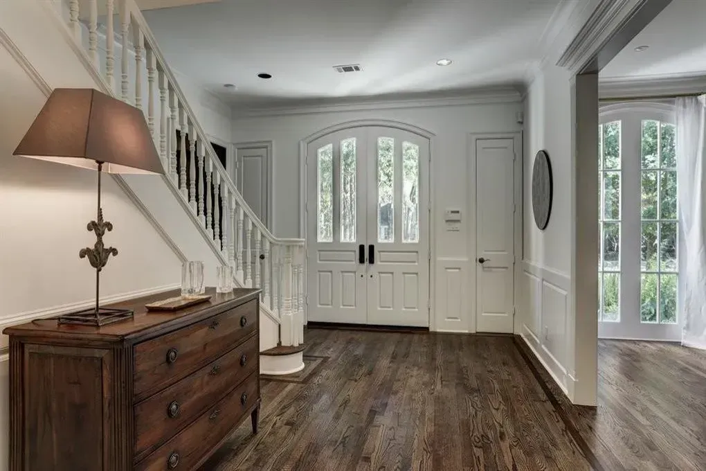 Entryway with wooden floors, staircase, and arched double doors. A dark wood dresser with lamp is on the left.