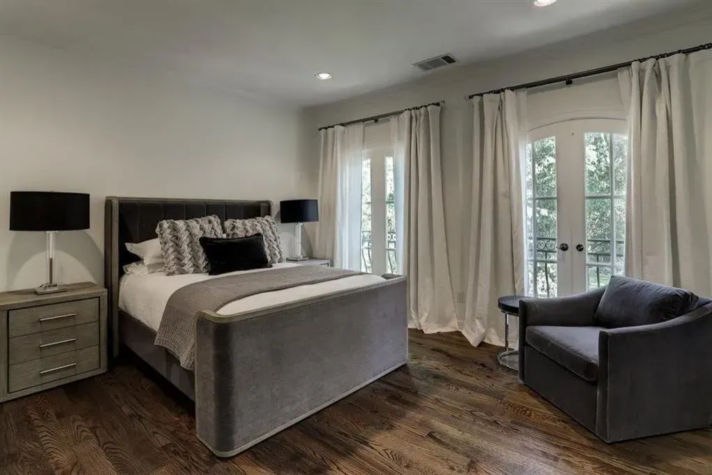 Bedroom with gray velvet bed, nightstands, arm chair, French doors, and dark wood floors.