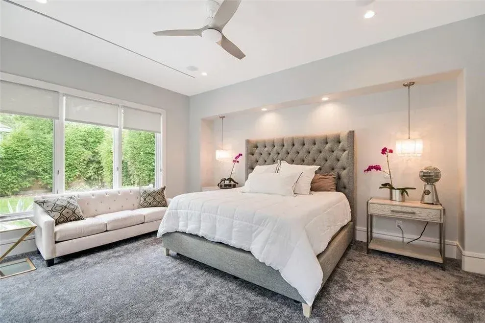 Gray bedroom with a tufted headboard bed, sofa, large window, and bedside table with lamps.