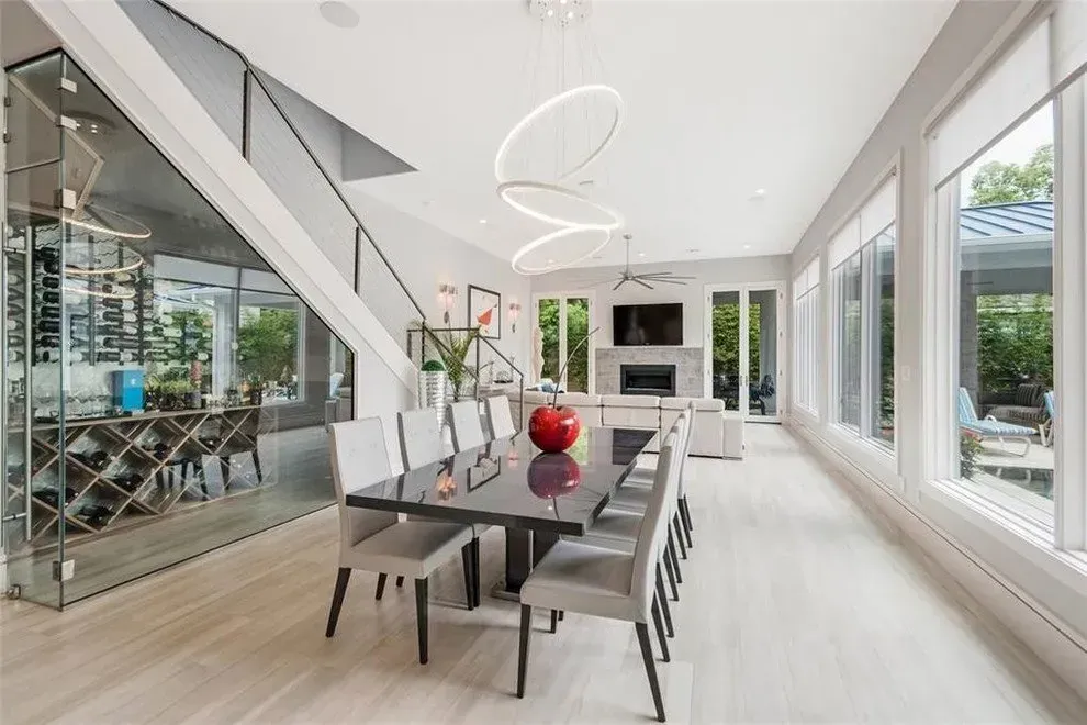 Modern dining room with a long table, chairs, and wine storage. Windows and a staircase are visible.