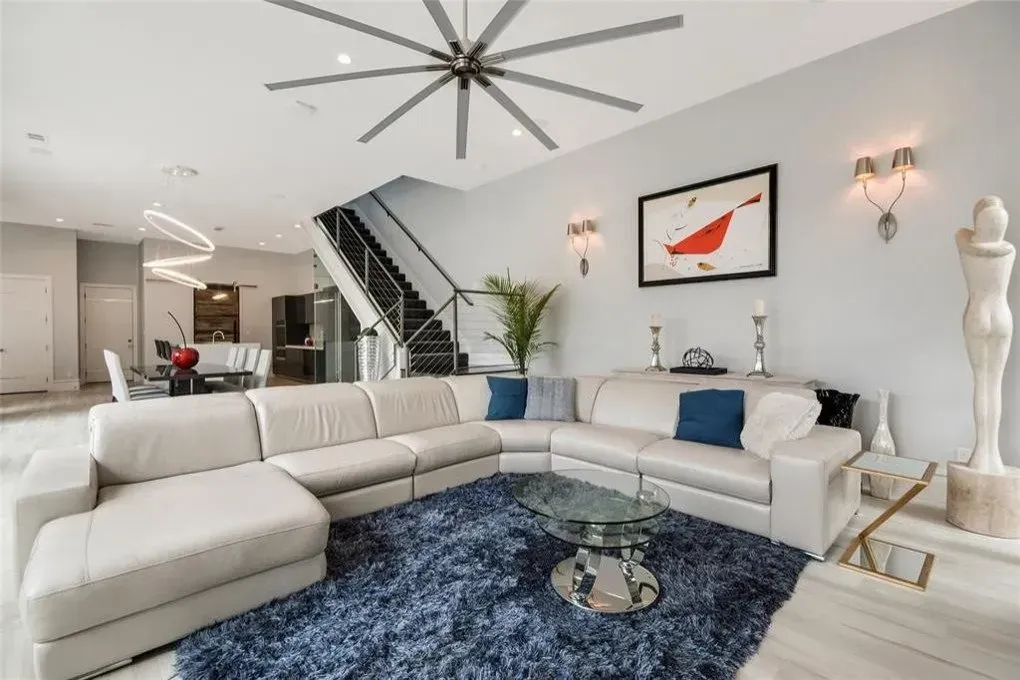 Modern living room with sectional sofa, blue rug, and artwork under a large ceiling fan.