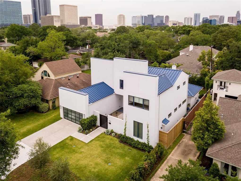 Modern white house with blue roof against a city skyline. Green lawn and trees surround it.