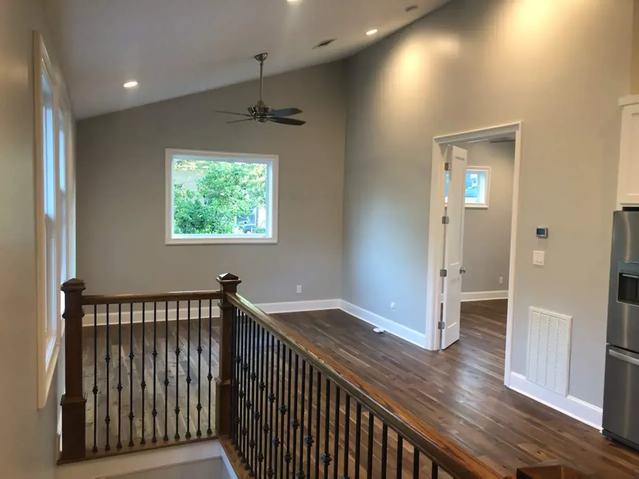 Interior with wood floors, railing, neutral walls, window, ceiling fan, and open doorway.