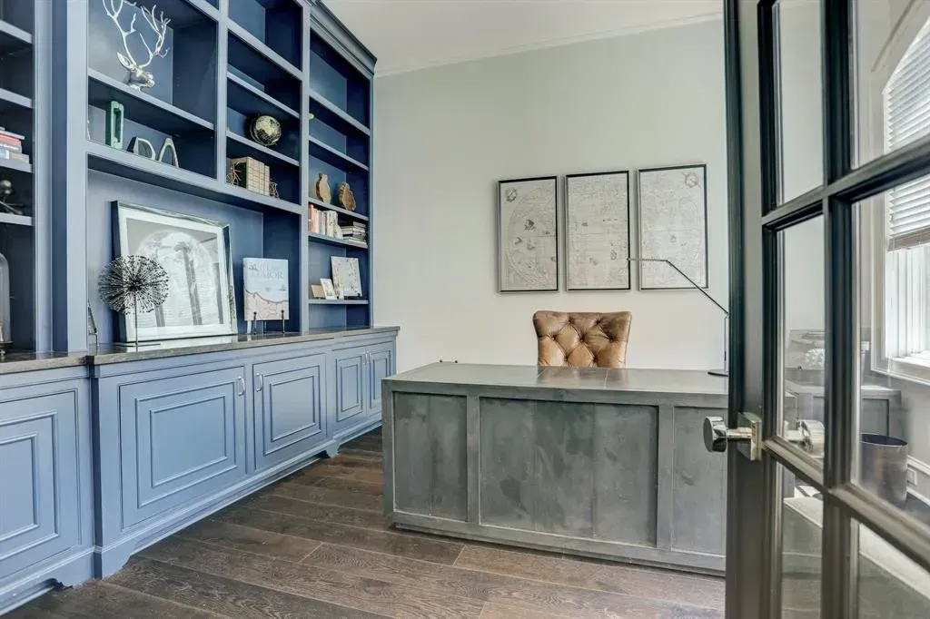 Blue home office with built-in bookshelves, gray desk, and tan tufted chair; light blue walls and wood floors.