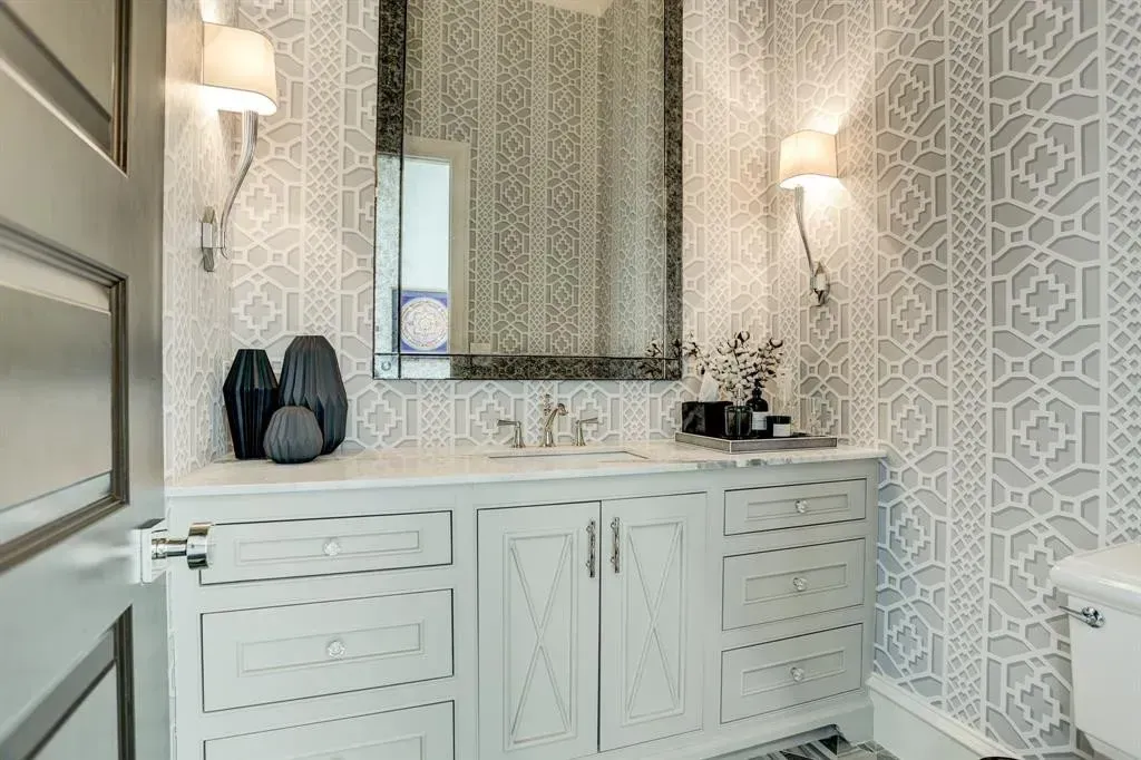 Bathroom with light gray and white patterned wallpaper, white vanity, mirror, and sconces.