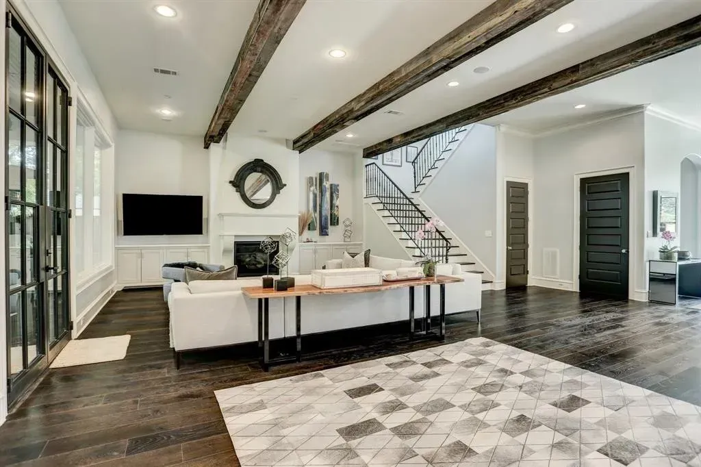 Spacious living room with white walls, dark wood floor, large rug, fireplace, and exposed beams.
