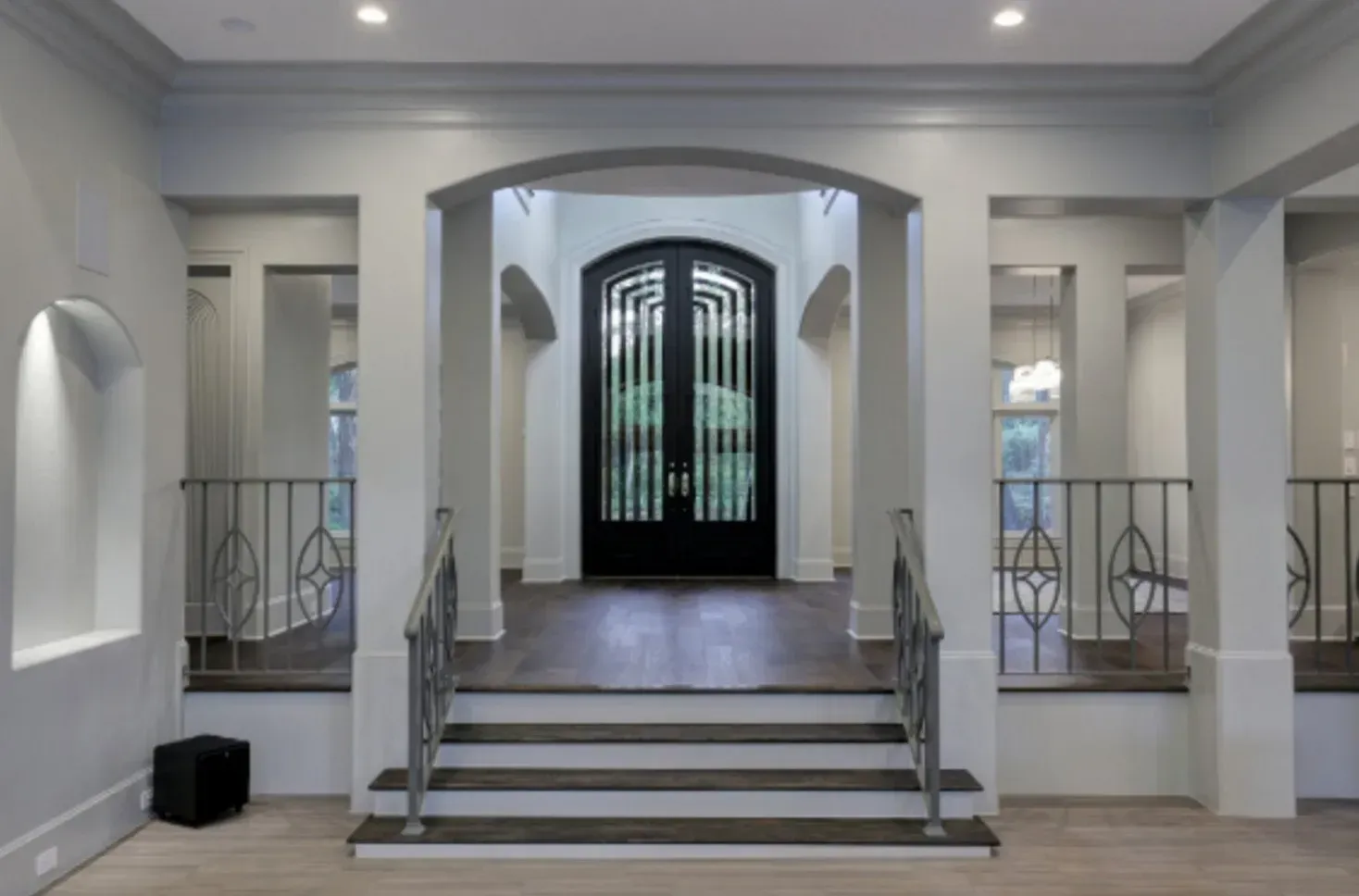 Grand entryway with dark front door, archway, and stairs. Light gray walls, dark wood floors.