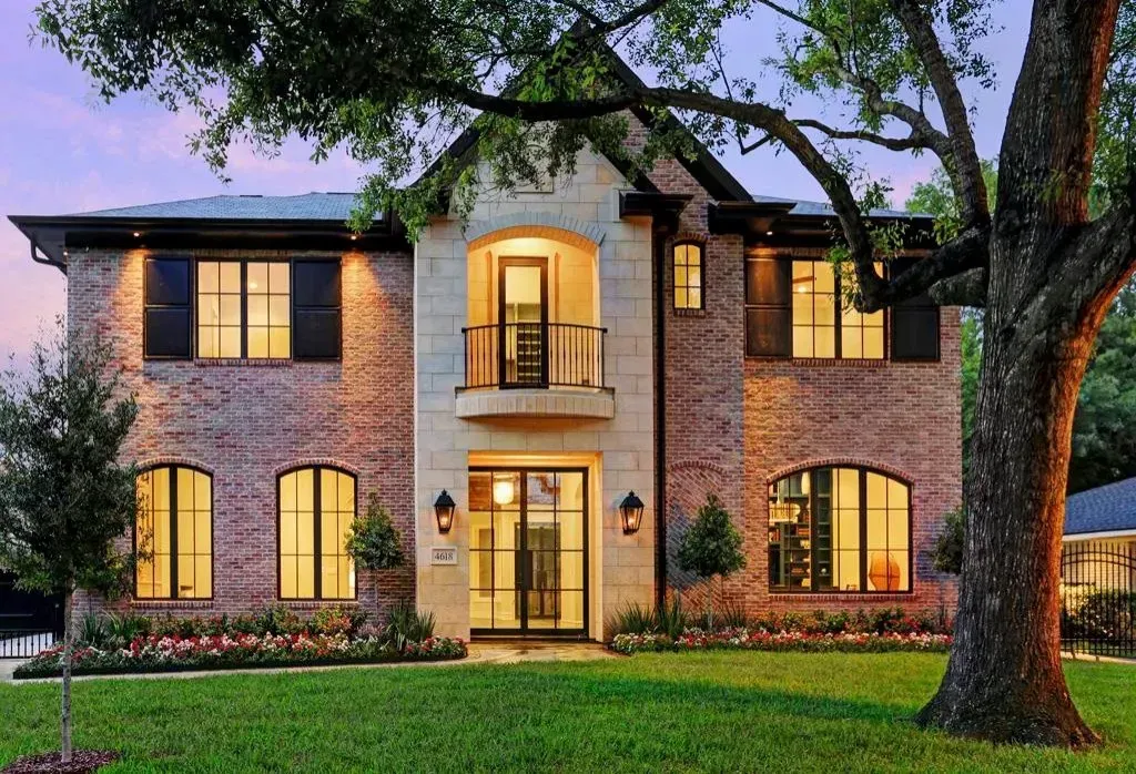 Two-story brick house with large windows, balcony, and lush green lawn at dusk.